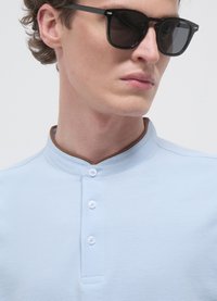 Polo azzurro chiaro con colletto marrone, dotata di abbottonatura e tessuto testurizzato; modello con occhiali da sole neri.