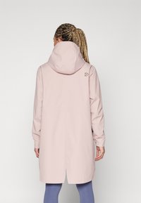 Imperméable rose clair avec une grande capuche, des manches longues et une coupe décontractée. Il comporte un petit logo à l'arrière du col et un ourlet légèrement arrondi.