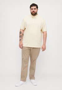 Cămașă polo dintr-un material galben deschis, cu mâneci scurte și guler, asociată cu pantaloni bej și adidași albi. Prezintă tatuaje pe braț.