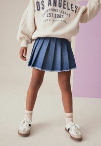 Denim plooirok in middelblauw met een rafelige zoom. Gepaard met een crèmekleurige oversized sweatshirt en witte sneakers met bruine accenten.