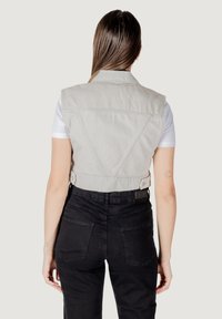 Gilet in denim beige tagliato con colletto strutturato e dettagli cuciti, indossato sopra una camicia bianca, abbinato a pantaloni neri a vita alta.