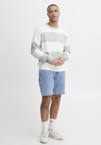 Fehér és világoskék csíkos pulóver, kerek nyakkal, kék denim rövidnadrággal. A modell fehér sportcipőt és bokazoknit visel, egy semleges környezetben áll.