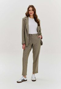 Kobieta stojąca w w kratkę marynarce i spodniach, w białej koszulce i białych trampkach, na jednolitym tle, z rękami w kieszeniach, uśmiechnięta.