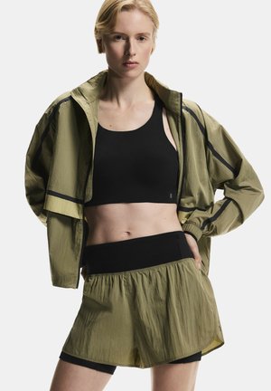 Športovo založená žena v olivovo zelenej ľahkej bunde a šortkách s čiernou športovou podprsenkou, stojaca s jednou rukou na bokoch pred jednofarebným pozadím.