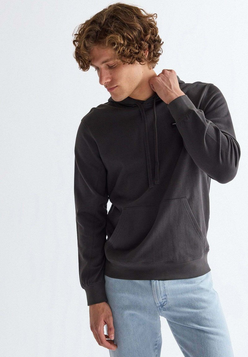 Junger Mann mit lockigem Haar, der einen dunkelgrauen Hoodie und hellblaue Jeans trägt, schaut nach unten und berührt mit einer Hand seinen Hals.