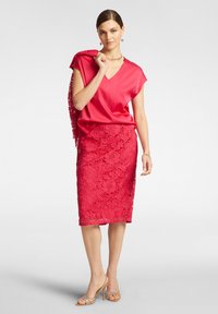 Blusa di raso rosso con maniche corte, abbinata a una gonna a tubino rossa in pizzo aderente, lunga fino al ginocchio, con motivi floreali. Gli accessori includono uno scialle.