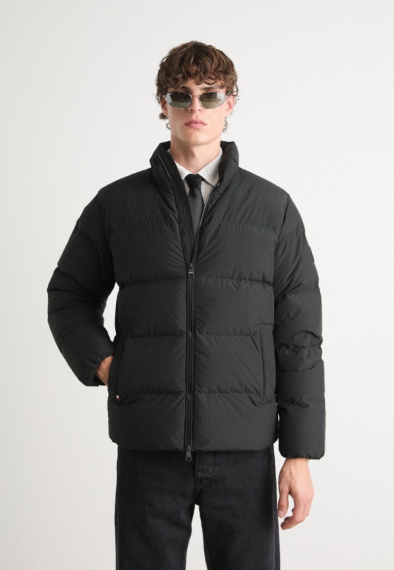 Jachetă puffer neagră, cu guler înalt, design matlasat orizontal, închidere cu fermoar și buzunare laterale. Purată peste o cămașă și o cravată.