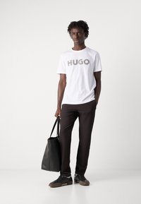 Hvit bomulls T-skjorte med "HUGO" logo i et mønster, kombinert med mørkebrune bukser og svart skinn tote bag, iført sorte sneakers.