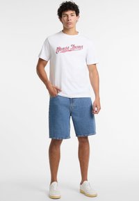 Camiseta blanca con el logo rojo de "Guess Jeans", combinada con shorts de mezclilla azul. El modelo lleva zapatillas blancas. Diseño simple, aspecto casual.