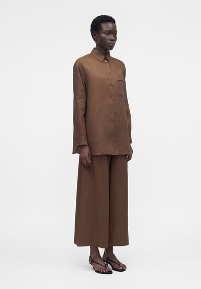 Ενήλικας που φοράει φαρδιά καφέ πουκάμισο με μακριά μανίκια και κουμπιά, παντελόνι με φαρδιά μπατζάκια που ταιριάζει και μαύρα σανδάλια με τακούνι, στέκεται μπροστά σε απλό ανοιχτό φόντο.