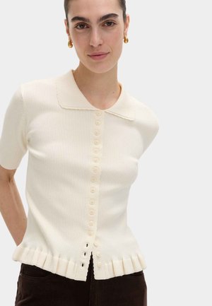 Vrouw draagt een kortemouwige crème ribgebreide top met kraag, verticale knopen aan de voorkant en een geplooide zoom, gecombineerd met donkere broek.