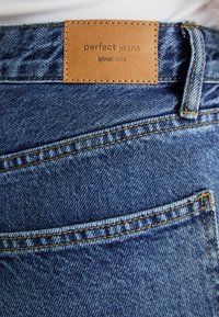 Blue denim jeans with a leather label reading "perfect jeans" and "ginatricot." Features yellow stitching and a smooth, textured surface.