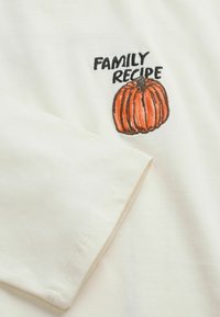 White fabric with a long sleeve and embroidered orange pumpkin next to black text reading "FAMILY RECIPE" near the shoulder area.