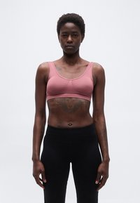 Ενήλικη γυναίκα με κοντά σγουρά μαλλιά, ντυμένη με ροζ αθλητικό σουτιέν και μαύρο κολάν, στέκεται με το πρόσωπο προς τα εμπρός μπροστά σε απλό φόντο.