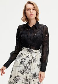 Blouse noire transparente avec des détails brodés et une fermeture boutonnée, associée à une jupe couleur crème à motif floral avec une taille froncée.