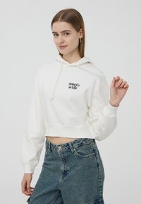 Felpa bianca corta con cappuccio regolabile e testo stampato "tranquilli" sulla parte anteriore; abbinata a jeans di jeans a vita alta.