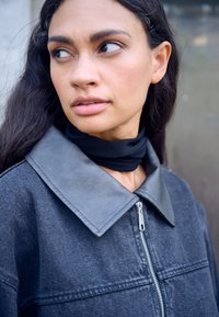 Kobieta o długich ciemnych włosach nosi ciemnoszarą kurtkę z dużym kołnierzem i czarny szalik, patrząc bokiem z neutralnym wyrazem twarzy.