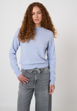 Junge Frau mit lockigem braunem Haar trägt einen hellblauen Pullover und hoch taillierte graue Jeans und steht mit einer Hand in der Hüfte.