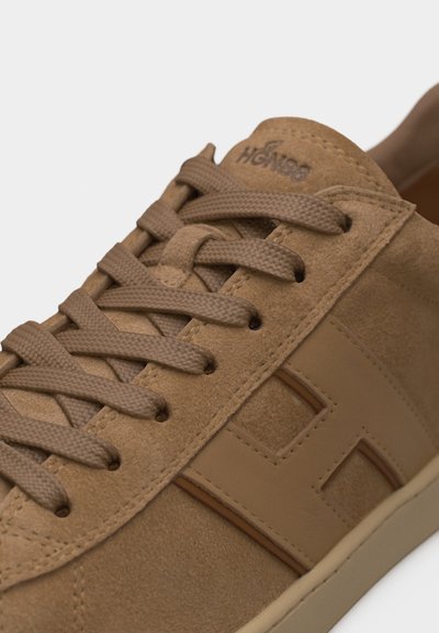 Κοντινό πλάνο ενός ταμπάνιου sneaker από σουέντ με κορδόνια στο ίδιο χρώμα και υφή λαστιχένιας σόλας, που έχει ένα μεγάλο ραμμένο "H" στη μία πλευρά.