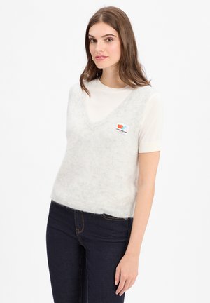 Jonge vrouw met bruin haar, gekleed in een witte top met korte mouwen en een lichtgrijze, fuzzy mouwloze sweater, over donkere jeans, staande tegen een witte achtergrond.