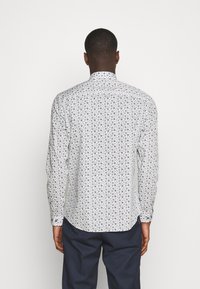 Biała koszula z długim rękawem z niebieskim okrągłym wzorem, z kołnierzykiem zapinanym na guziki i zaokrąglonym hemem. Materiał wydaje się lekki i gładki.