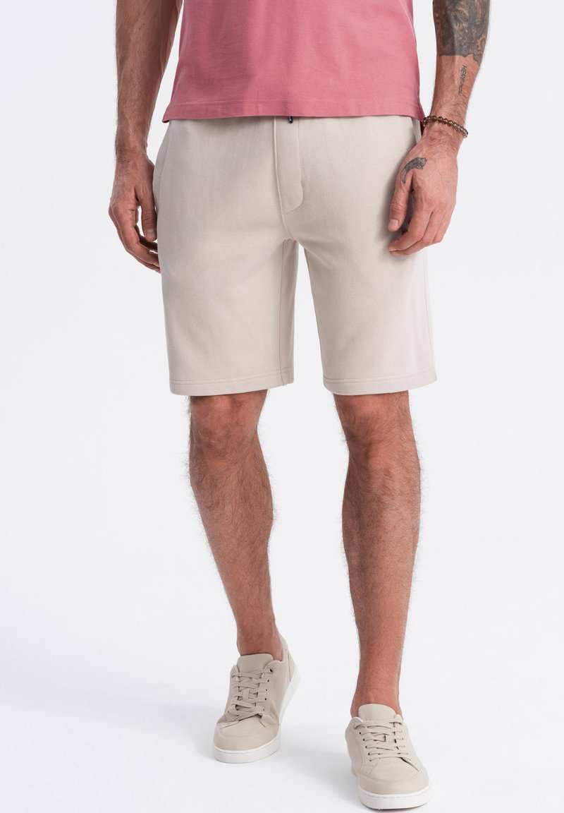 Shorts beige en tissu mélangé coton, dotés d'une taille élastique et d'une texture lisse. Associés à des baskets de couleur claire.