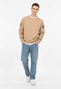 Felpa beige con diverse stampe grafiche colorate sulle maniche, abbinata a jeans in denim azzurro chiaro e sneaker bianche. Tessuto morbido e vestibilità casual.