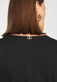 Femme aux cheveux bruns raides portant un haut noir avec un petit logo en or Twinset au niveau de l'encolure arrière et une boucle d'oreille en or.