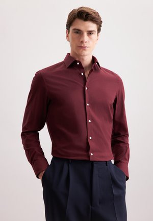 Chemise rouge à manches longues avec col boutonné, ornée de boutons blancs et à coupe ajustée. Associée à un pantalon bleu marine.