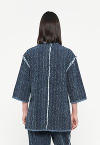 Blue textured garment with a loose fit, wide sleeves, and frayed edges. Features a vertical seam down the back for added detail.
