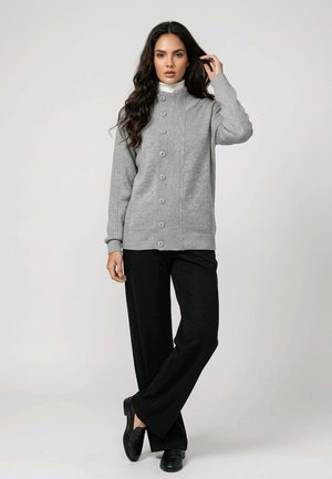Mujer con un cárdigan gris abotonado, pantalones negros de pierna ancha y zapatos negros, de pie con un pie cruzado delante del otro.