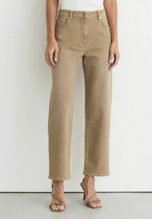 Vrouw draagt beige jeans met hoge taille en rechte pijpen, met vijf zakken en bruine open-toe sandalen met hak, staand tegen een witte muur.
