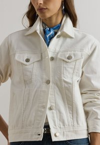 Balta denima jaka ar klasisku dizainu, kurai ir divas krūšu kabatas, sudraba detaļas un pamanāmi šuvju elementi.