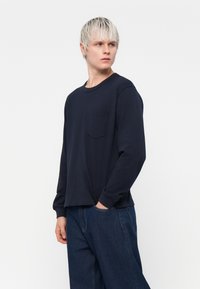 Merinokangas pitkähihaisessa navy-paidassa, jossa on pyöreä kaula-aukko ja rintatasku. Yhdistetty löysiin farkkulleihin.