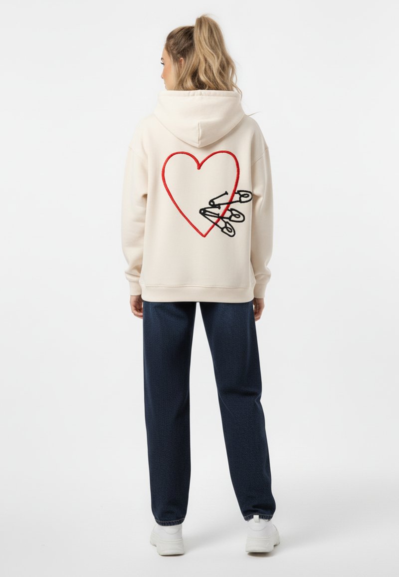 Felpa crema con design di cuore rosso e spilloni neri sulla schiena. Abbinata a pantaloni blu texture e sneakers bianche.