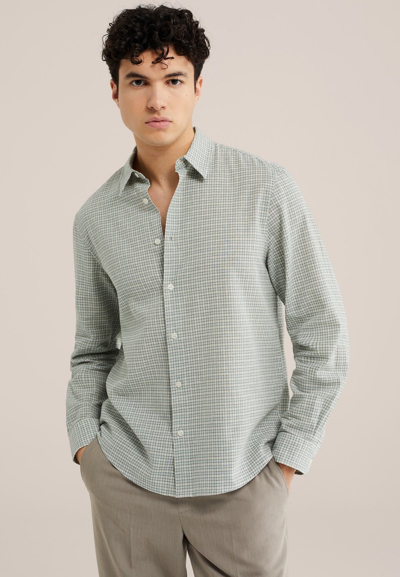 Jeune homme aux cheveux bouclés foncés portant une chemise à boutons à carreaux verts clairs et blancs et un pantalon beige, mains dans les poches, fond neutre.
