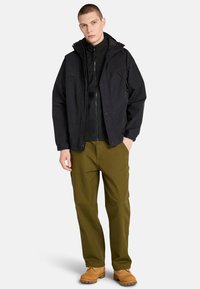 Musta ulkotakki jossa on huppu, mustan vetoketjullisen sisätakin päällä. Oliivinvihreät housut ja vaaleat saappaat täydentävät asukokonaisuuden.