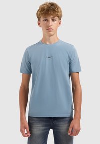 Tienerjongen met lichtbruin haar, gekleed in een lichtblauw T-shirt met korte mouwen en vervaagde blauwe jeans, staande tegen een effen grijze achtergrond.