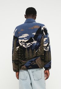 Jachetă din fleece cu un model de peisaj montan în albastru, alb și verde. Croială supradimensionată, cu guler și țesătură texturată.