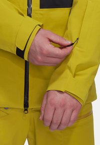 Gelbe wasserdichte Jacke mit einem schwarzen Reißverschluss, die eine Nahaufnahme eines Ärmels mit Velcro-Verschluss und einer Reißverschlusstasche auf dem Ärmel zeigt.