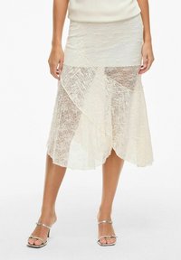 Jupe midi en dentelle de couleur blanc cassé, présentant des motifs texturés et des panneaux asymétriques. Fabriquée en matériau léger et transparent. Sandales argentées incluses.
