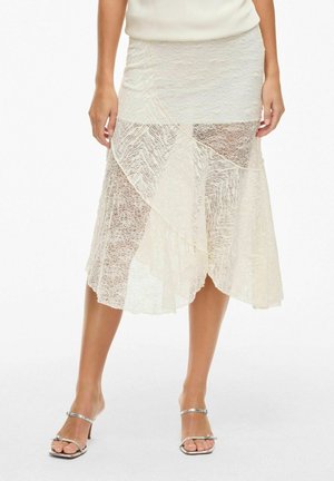Lace midi skirt in off-white, featuring textured patterns and asymmetrical panels. Made of lightweight, sheer material. Silver sandals included.