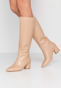 Nude knee-high boots made of smooth leather, featuring a pointed toe and a chunky heel, with a fitted shaft and no visible hardware.