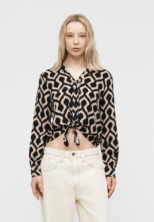 Γυναίκα με ίσια ξανθά μαλλιά φοράει μακρυμάνικο πουκάμισο σε μαύρο και μπεζ σχέδιο, δεμένο στη μέση, και ψηλόμεσο εκρού παντελόνι.