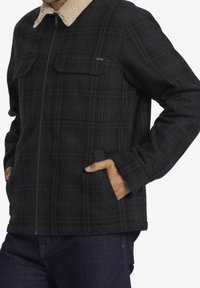 Plaid jacket made of black and dark green fabric, featuring a beige sherpa collar, two chest pockets, and a zip closure.
