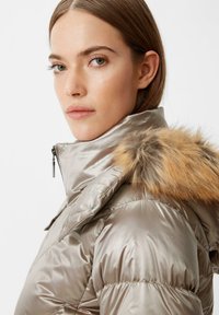 Metaliczna beżowa kurtka puchowa z kapturem wykończonym futrem, z wysokim kołnierzem i zapięciem na zamek. Teksturowana tkanina o błyszczącym wykończeniu.