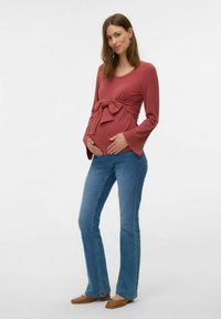 Top da maternità color ruggine, in tessuto a coste con un nodo in vita, maniche lunghe; abbinato a jeans a gamba larga blu e scarpe basse marroni.
