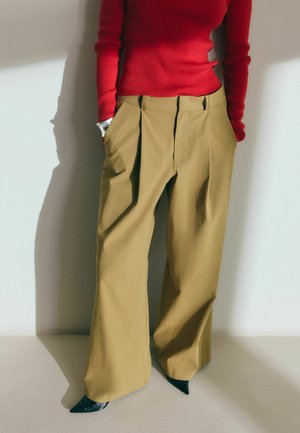 Person iført vidde bukser i khaki, rød tætsiddende ribstrikket sweater og sorte spidse sko, stående med hænderne i lommerne.