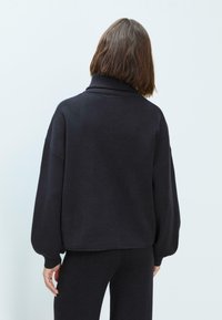 Pull en maille noir avec un col montant, coupe oversize et manches ballons. Texture douce, poignets et ourlet côtelés. Design simple sans motifs.