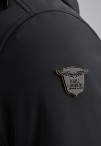 Chaqueta negra con un distintivo texturizado que presenta alas y el texto "PME LEGEND AMERICAN CLASSIC", destacando su diseño elegante y material.